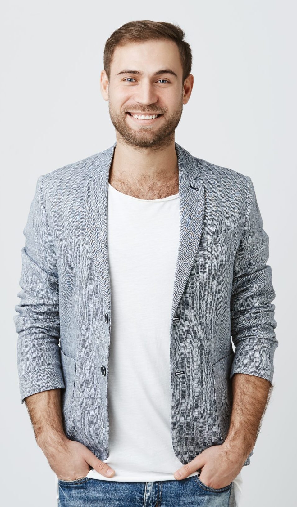 Delightful european man wearing jacket over white t-shirt and jeans, looks pleasantly at camera, has good mood as comes home after work. Fashionable pleasant looking male entrepreneur poses against gray studio wall